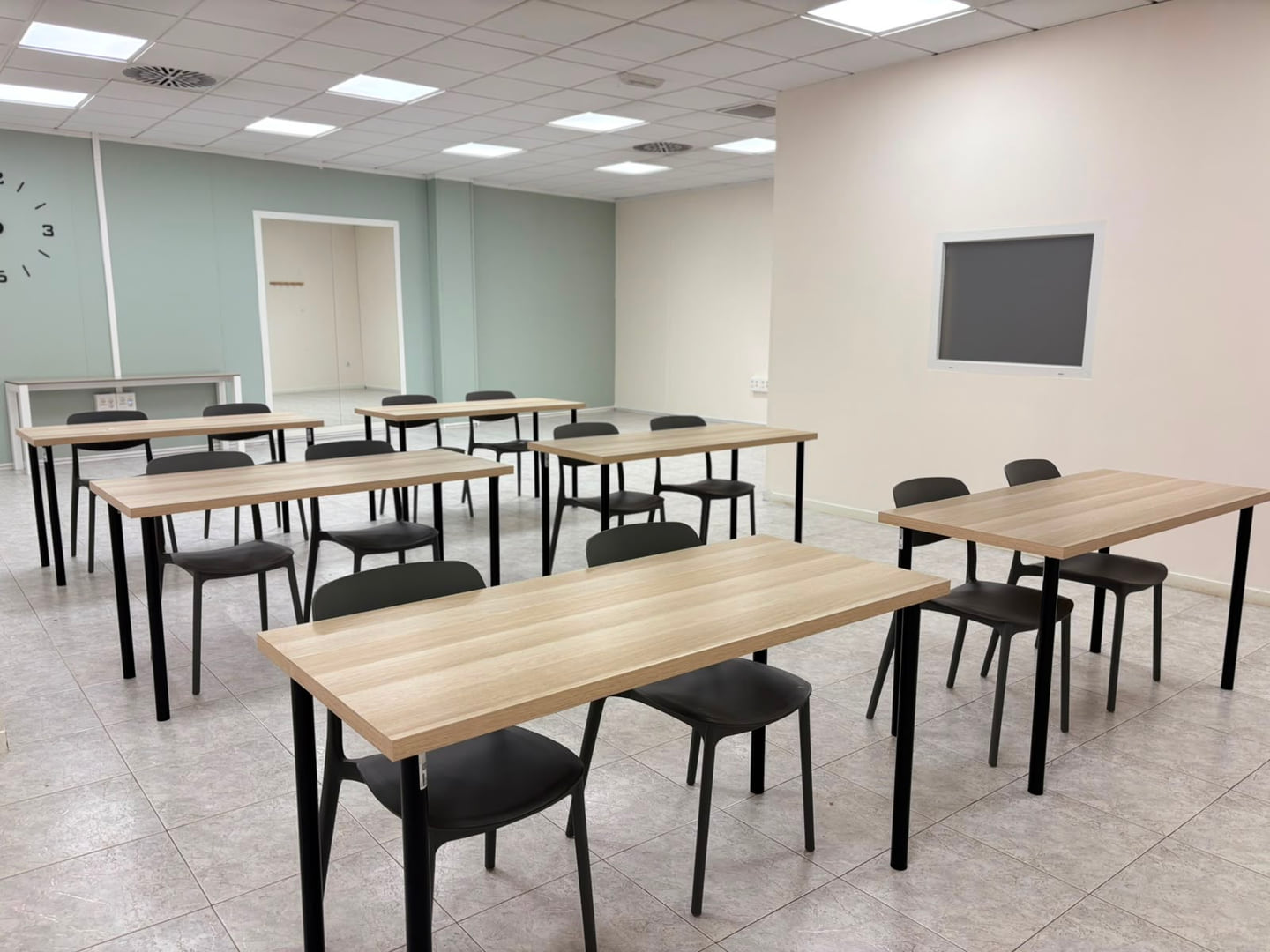 Mesas rectangulares de melamina clara con sillas negras, colocadas en filas frente a una pantalla de pared, bajo la luz uniforme del techo
